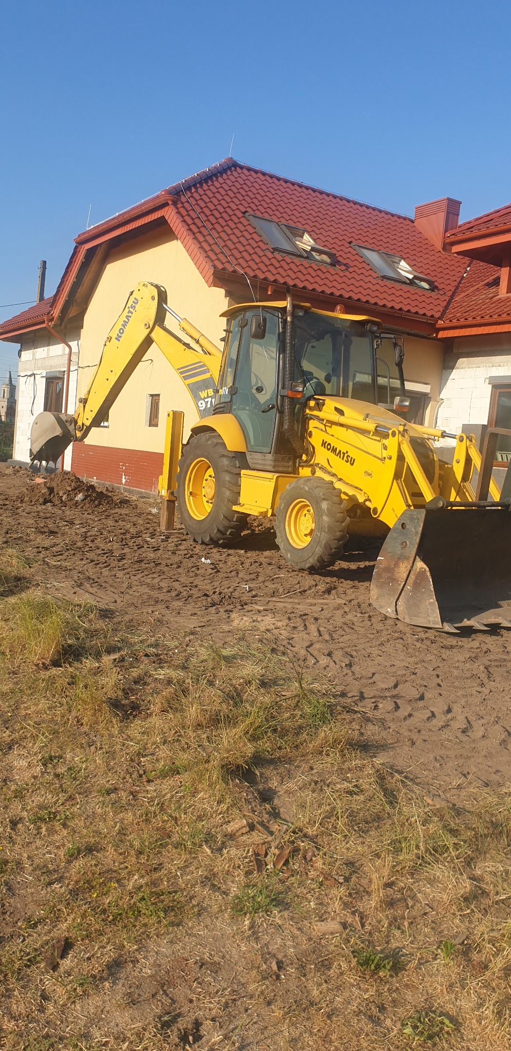Żółta koparko-ładowarka Komatsu WB 97S pracuje na placu budowy, obok domu z czerwoną dachówką i widocznym oknem w stanie surowym.