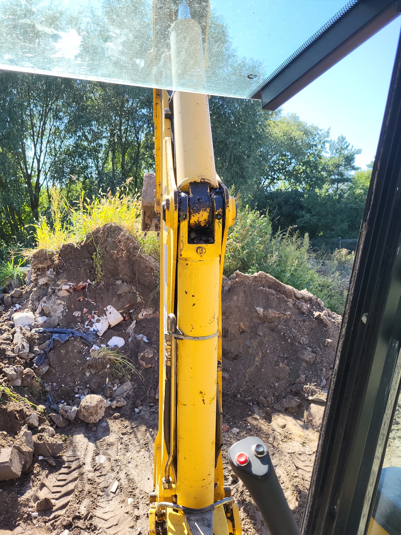 Widok z kabiny koparki na wykopaną ziemię z gruzem, zarys drzew w tle.