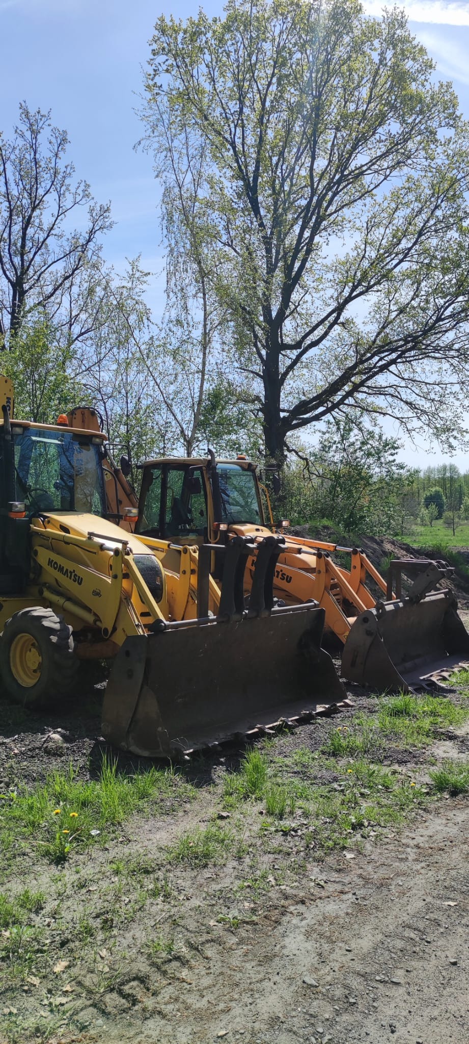 Dwie żółte koparko-ładowarki Komatsu zaparkowane na terenie budowy z widocznymi śladami opon na ziemi i drzewami w tle, uchwycone w słoneczny dzień.