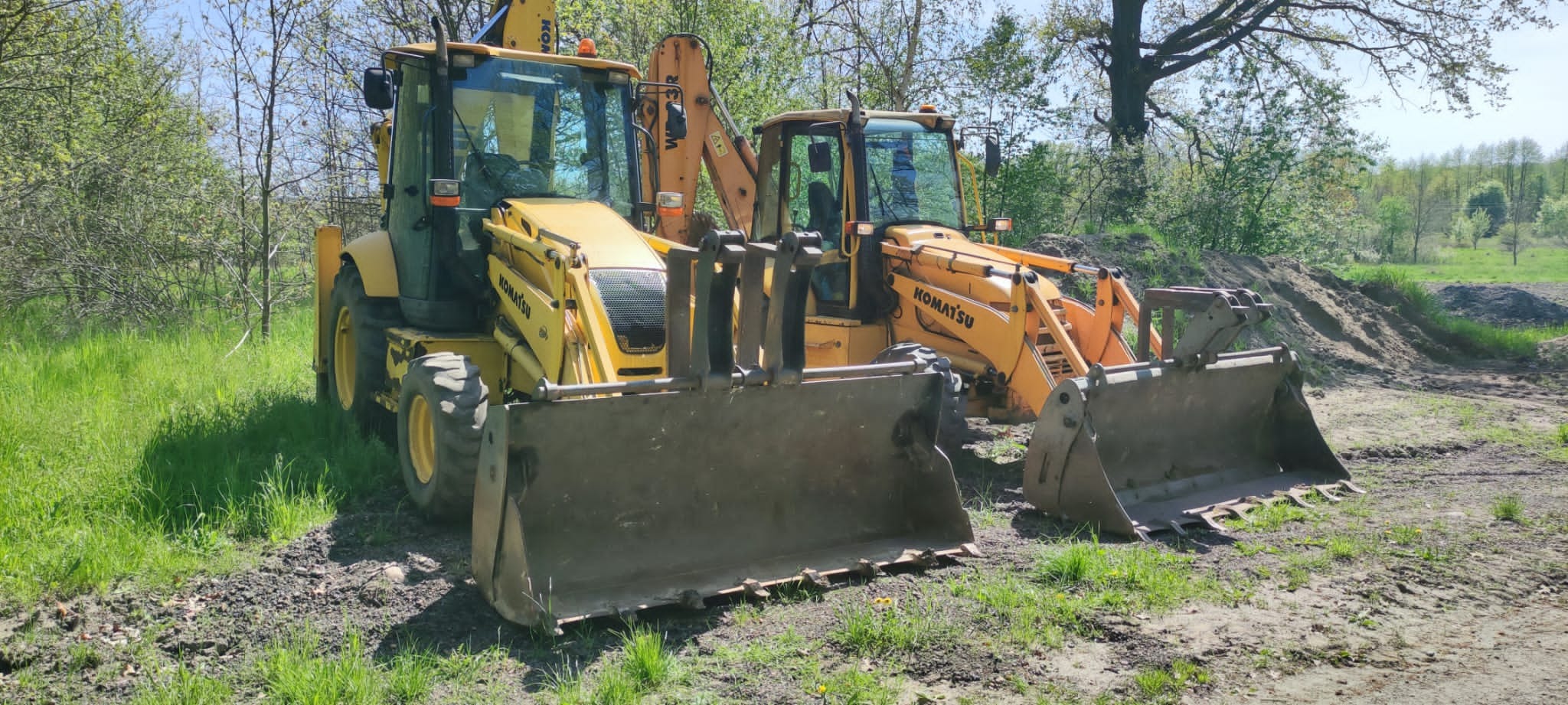 Dwie żółte koparko-ładowarki Komatsu stojące obok siebie na placu budowy z pryzmą ziemi w tle.