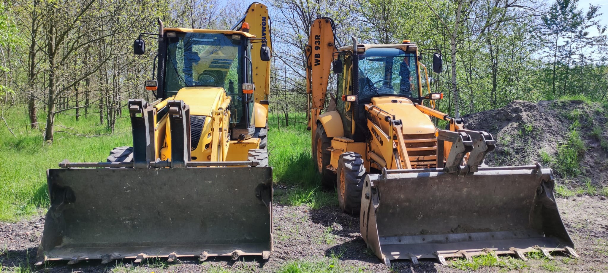 Dwie żółte koparko-ładowarki Komatsu WB93R stoją obok siebie na trawiastym terenie, z widoczną stertą ziemi w tle i drzewami bez liści w tle.
