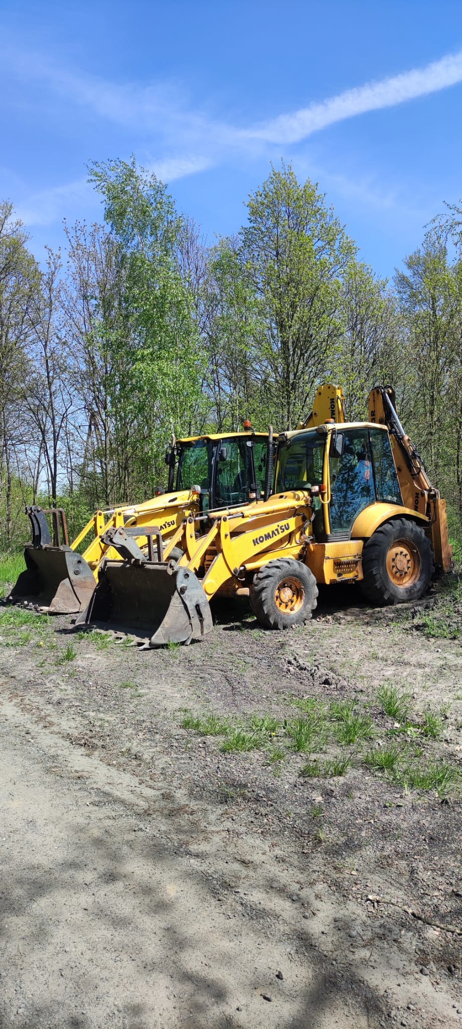 Dwie żółte koparko-ładowarki Komatsu stojące na nierównym, piaszczystym terenie z młodą trawą, w tle drzewa i błękitne niebo.