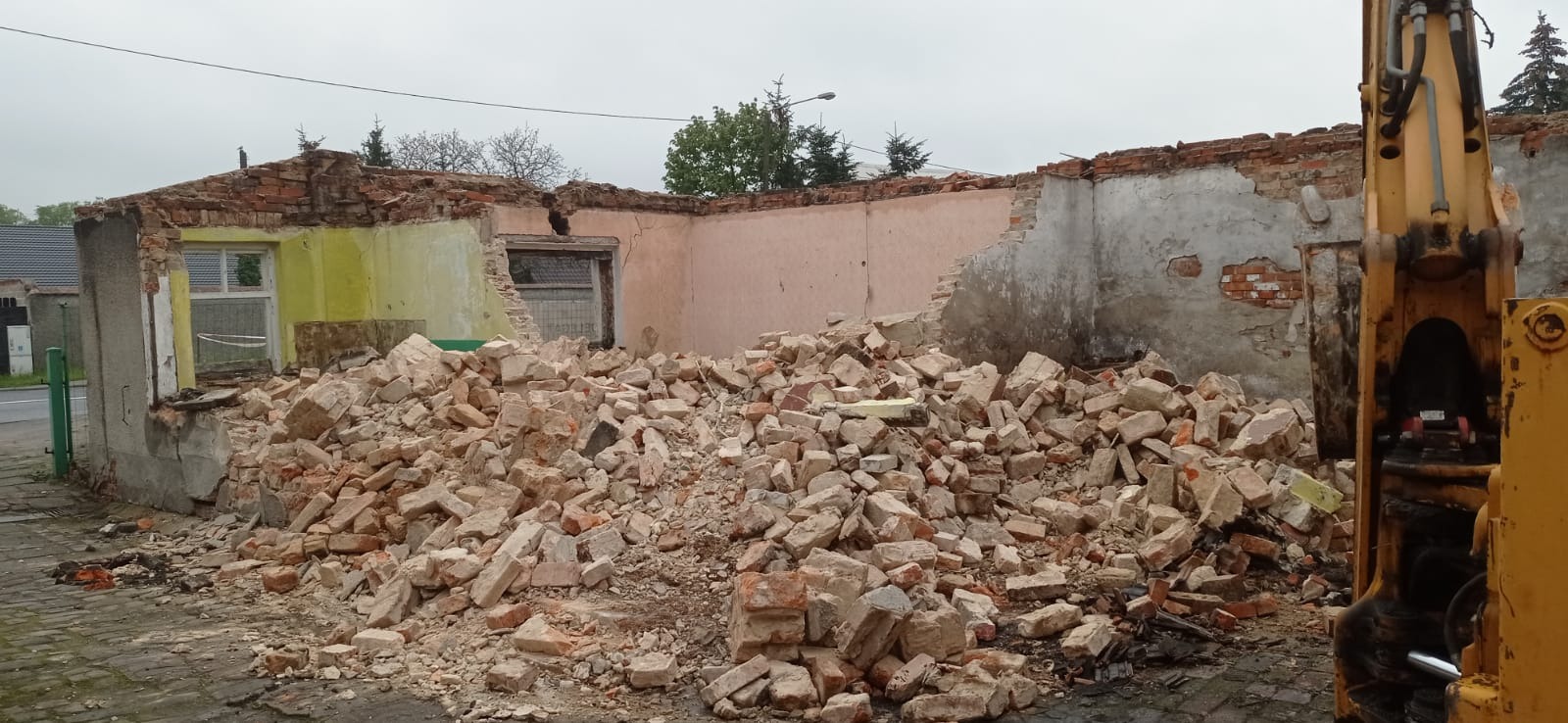 Sterta gruzu ceglanego przed częściowo zburzonym budynkiem z żółtą i różową ścianą, widoczny fragment żółtej koparki po prawej stronie kadru.