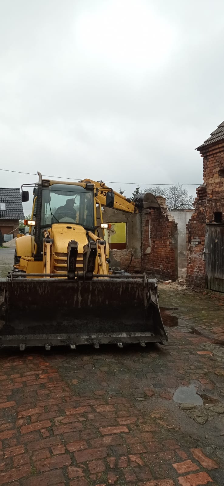 Żółta koparko-ładowarka z podniesioną łyżką przed częściowo zburzonym ceglanym budynkiem, widoczne cegły i gruz, na pierwszym planie mokra, ceglana nawierzchnia.