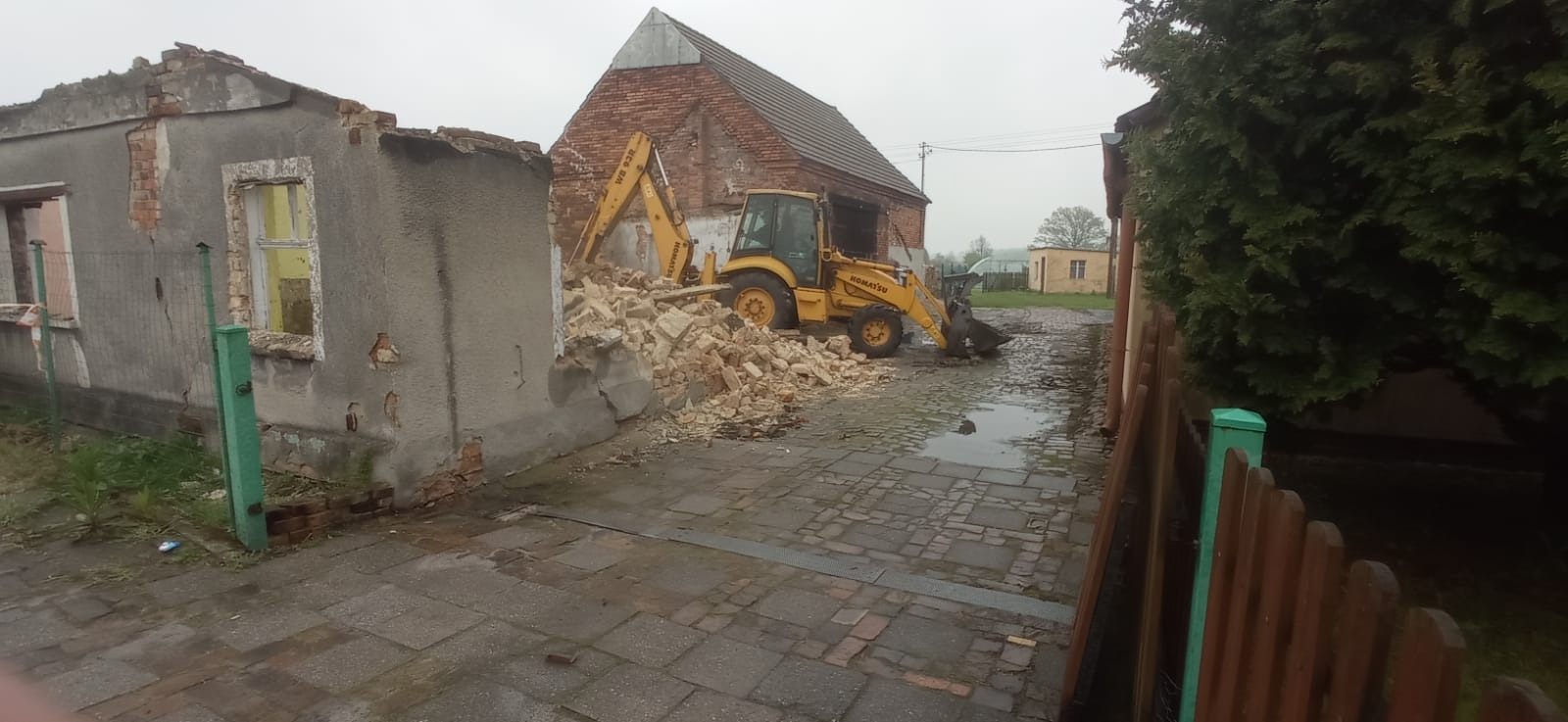 Żółta koparko-ładowarka Komatsu przy częściowo zburzonym budynku, gruz na brukowanej drodze, w tle budynek gospodarczy z czerwonej cegły.