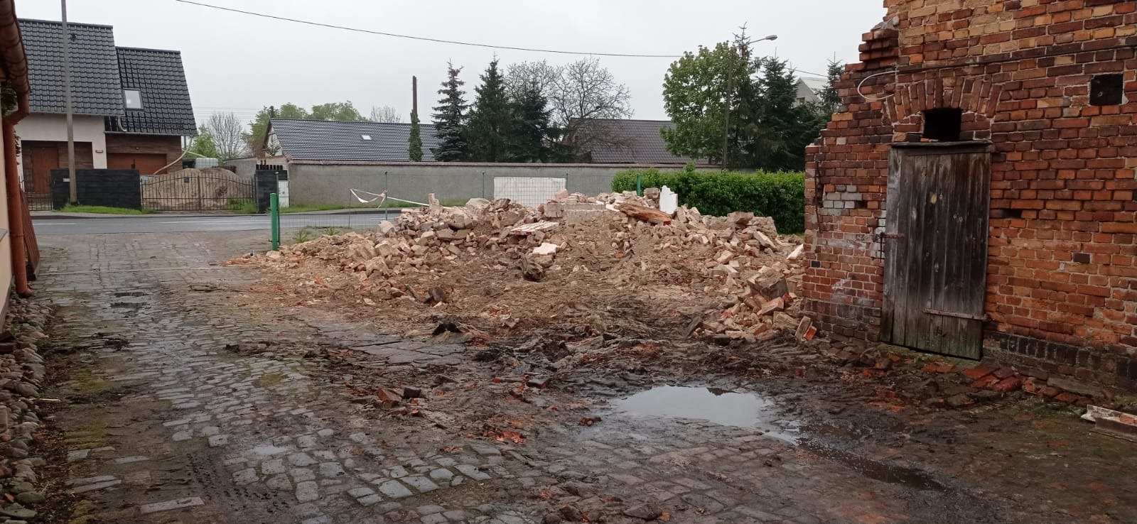 Sterta gruzu ceglanego po wyburzeniu ściany, widoczna brama w murze z cegły, mokra brukowa nawierzchnia, w tle zabudowania mieszkalne.