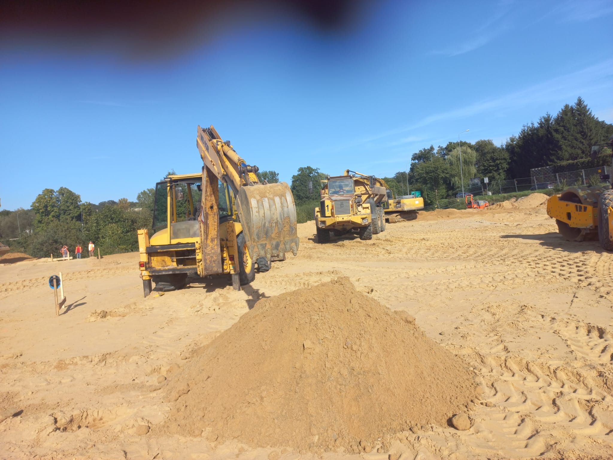 Żółta koparka z łyżką i walec na piaszczystym terenie budowy pod błękitnym niebem; w tle widoczne drzewa i pracownicy w odblaskowych kamizelkach.