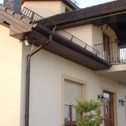 Jasna elewacja domu z brązowymi elementami wykończeniowymi, balkon z kutą balustradą, widoczny fragment dachu i rynny, oświetlenie zewnętrzne, flaga Polski powiewająca na balkonie.