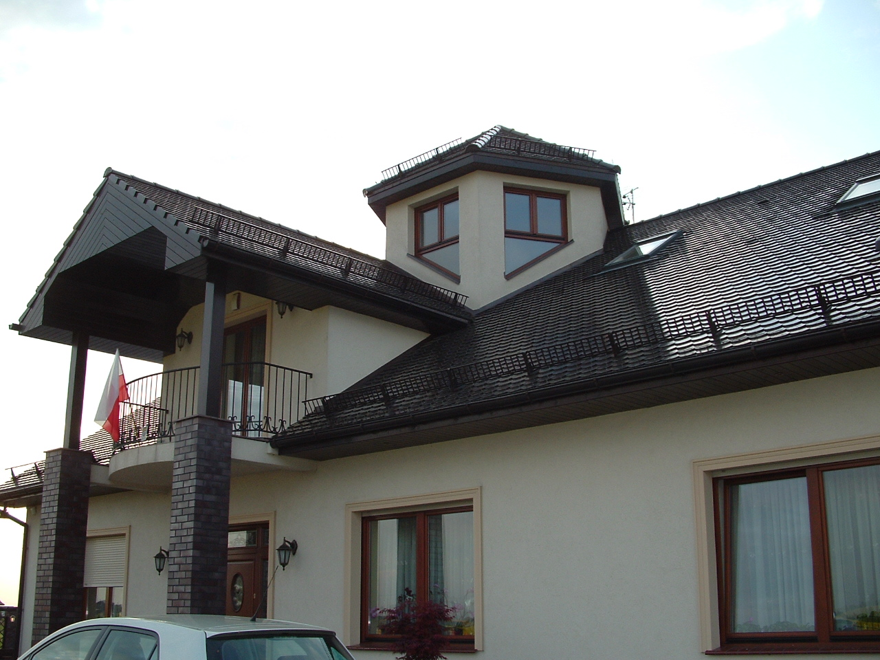 Dwukondygnacyjny dom z beżową elewacją, ciemnobrązowym dachem z dachówki ceramicznej i balkonem z flagą Polski, widoczny fragment samochodu.