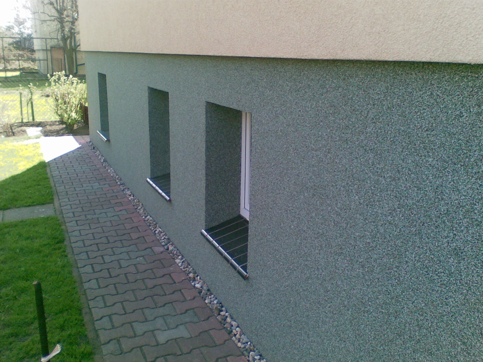 Fragment budynku z ocieploną elewacją w odcieniach szarości i beżu, widoczne trzy okna piwniczne z kratami, obok chodnik z kostki brukowej i trawnik.