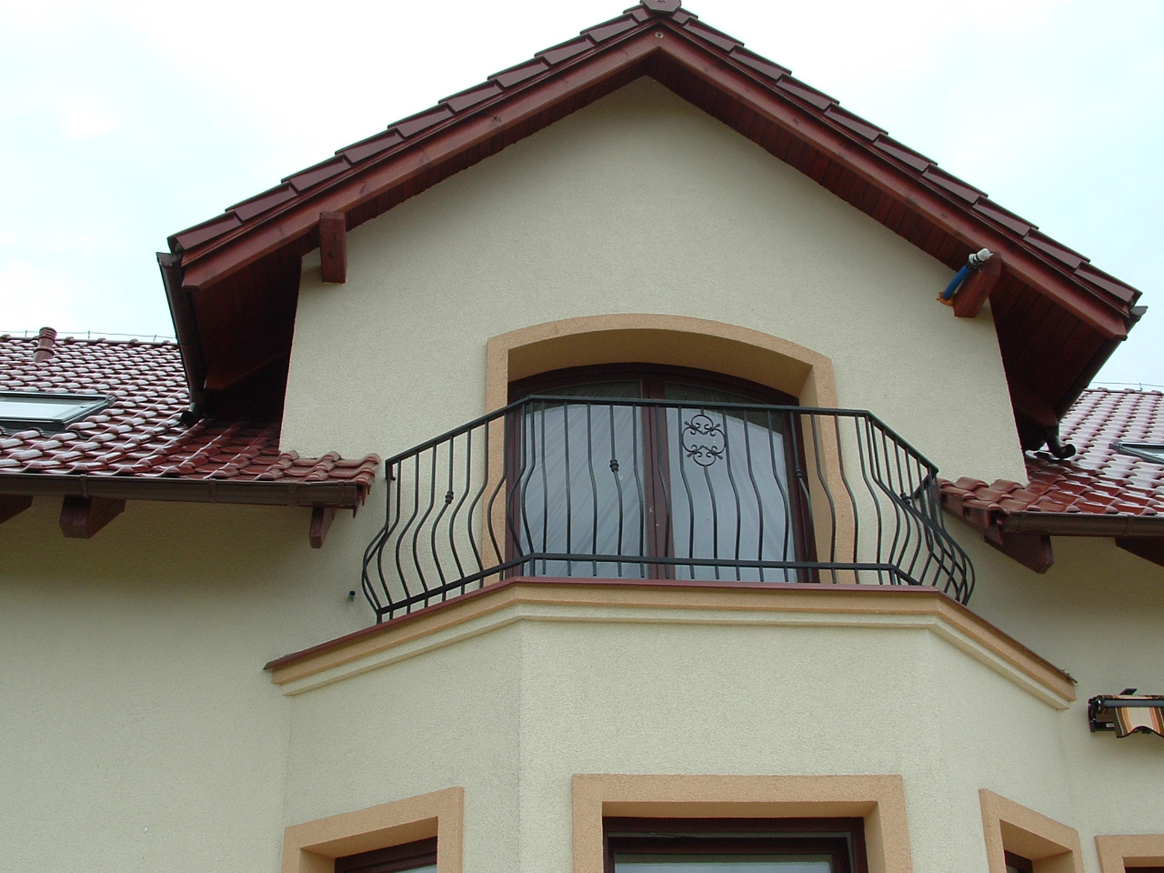 Fragment fasady domu z balkonem, ocieplona beżowa elewacja z dekoracyjnymi obramowaniami okien i balkonu, czerwona dachówka, widoczny fragment orynnowania.