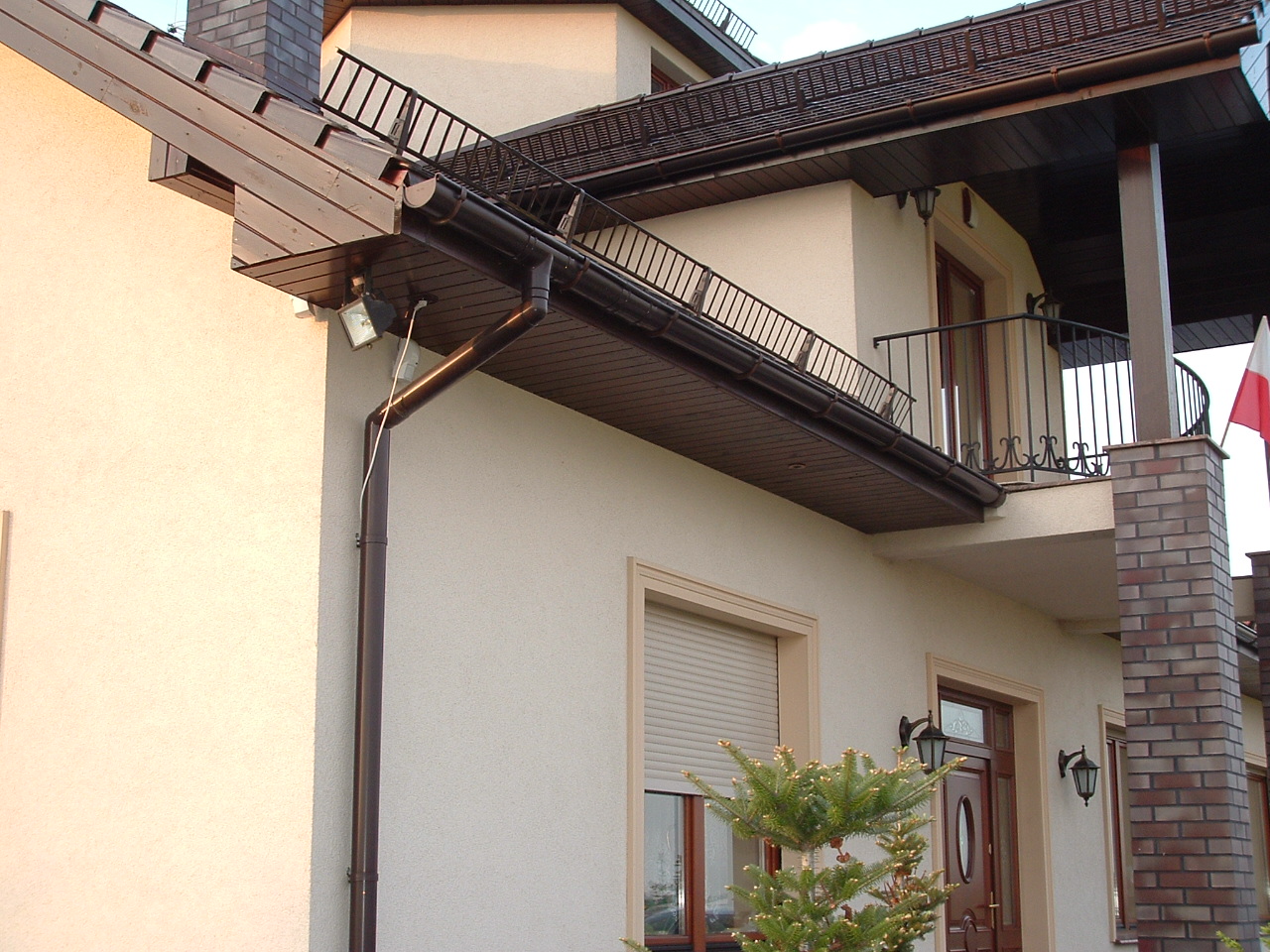 Jasna elewacja domu z brązowymi elementami wykończeniowymi, balkon z kutą balustradą, widoczny fragment dachu i rynny, oświetlenie zewnętrzne, flaga Polski powiewająca na balkonie.