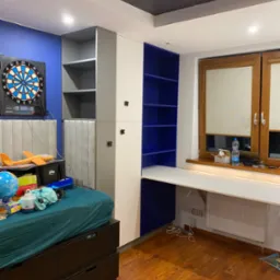 Pokój młodzieżowy z zabudową meblową: szafa z szarymi i białymi frontami, niebieskie półki, białe biurko pod oknem z drewnianą ramą, ciemna podłoga, sufit z motywem kosmosu, tarcza do darta...