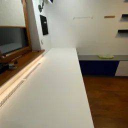 Długi, biały blat roboczy zamontowany wzdłuż ściany, z widocznym grzejnikiem pod oknem i lampką biurkową. Na ścianie wiszą półki i dekoracje.