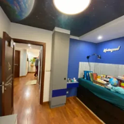 Pokój młodzieżowy z niebieską ścianą, biurkiem, łóżkiem z zagłówkiem i szafą w kolorze szarym i niebieskim. Na suficie fototapeta z motywem kosmosu i planety. Widoczny fragment korytarza...