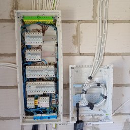 Ma-Gi Mariusz Gilewski Firma Handlowo-Usługowa - Dwie rozdzielnice elektryczne na ścianie z jasnych pustaków, jedna z kompletnym okablowaniem i rzędami bezpieczników, druga z nawiniętymi białymi kablami. Multimetr wisi obok, a kable są schludnie...