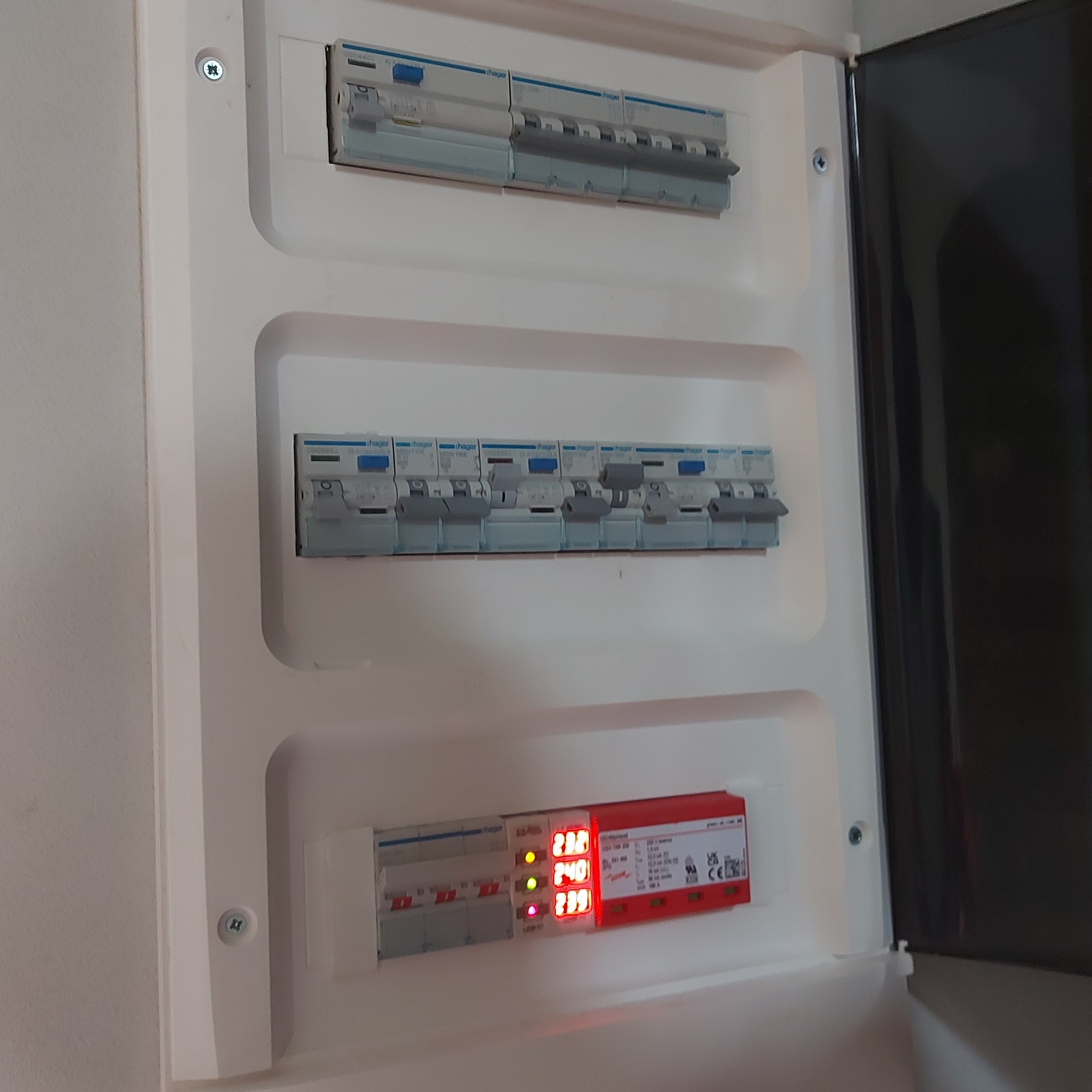 Rozdzielnica elektryczna z otwartymi drzwiczkami, widoczne zabezpieczenia i wyświetlacz napięcia. Estetyczne wykonanie, biała obudowa wkomponowana w ścianę.