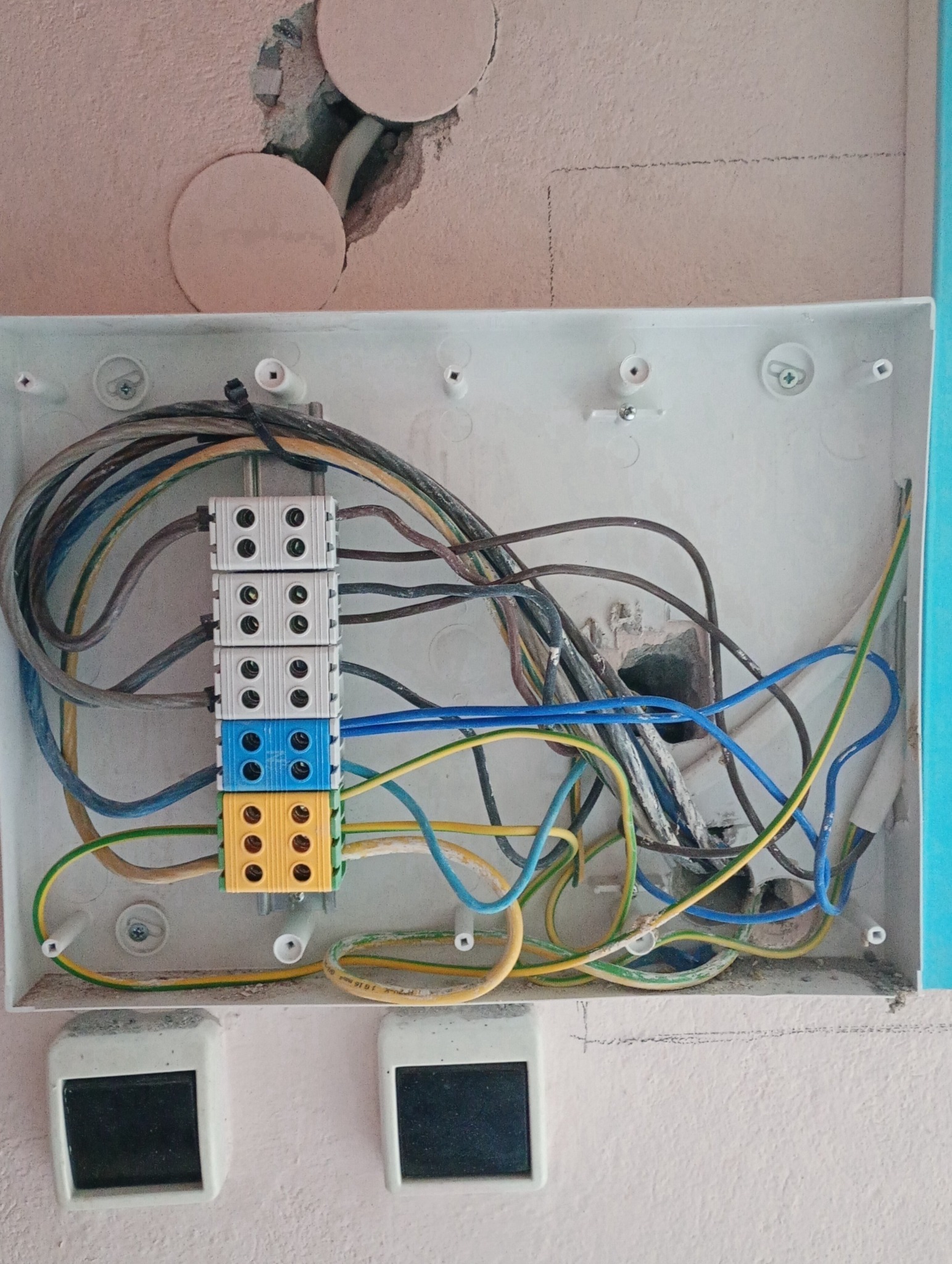 Otwarta puszka elektryczna z widocznymi przewodami i złączkami w kolorach: białym, niebieskim i żółtym. Nad puszką widoczne otwory w ścianie.
