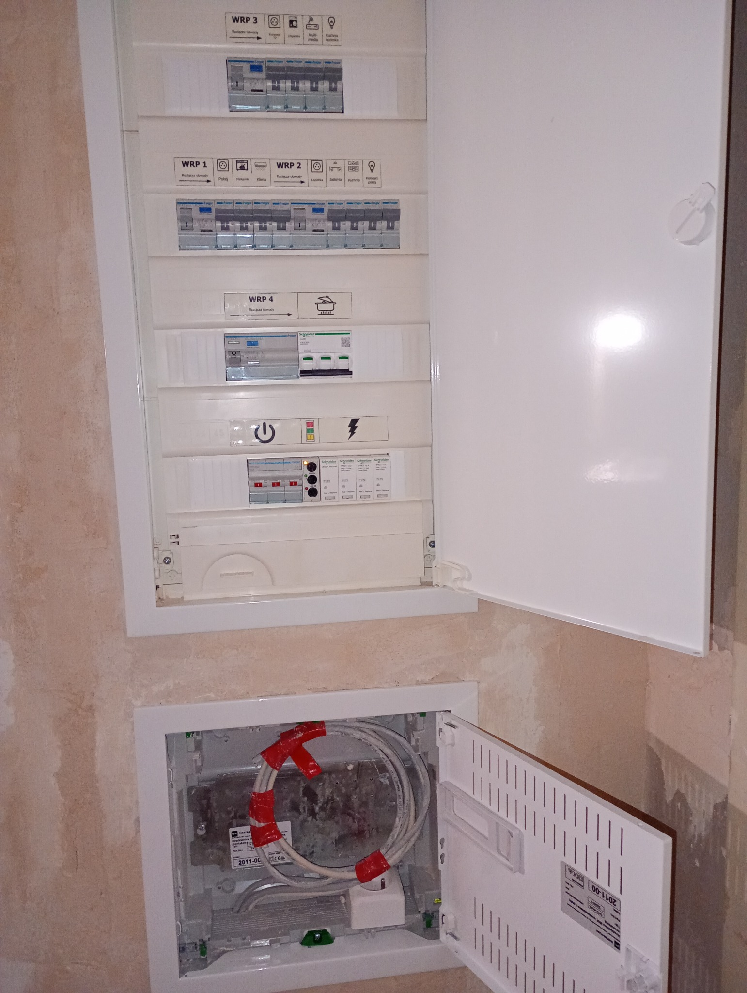 Otwarta skrzynka rozdzielcza z bezpiecznikami i okablowaniem w ścianie. Widoczne oznaczenia obwodów i zabezpieczenia. Dolna skrzynka z kablami światłowodowymi.