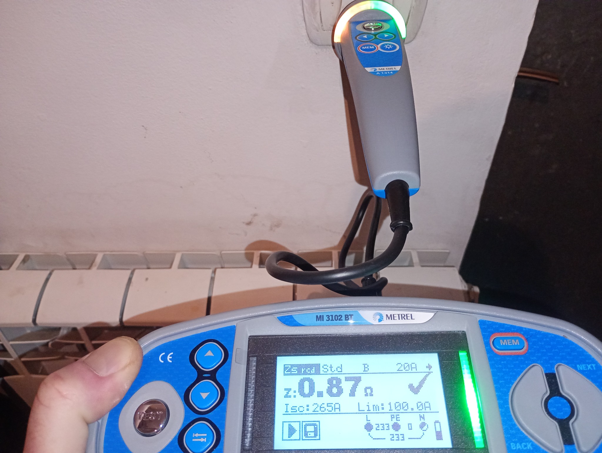 Miernik Metrel MI 3102 BT podczas pomiaru gniazdka elektrycznego. Wyświetlacz pokazuje wynik pomiaru impedancji pętli zwarcia. Widoczny fragment grzejnika.