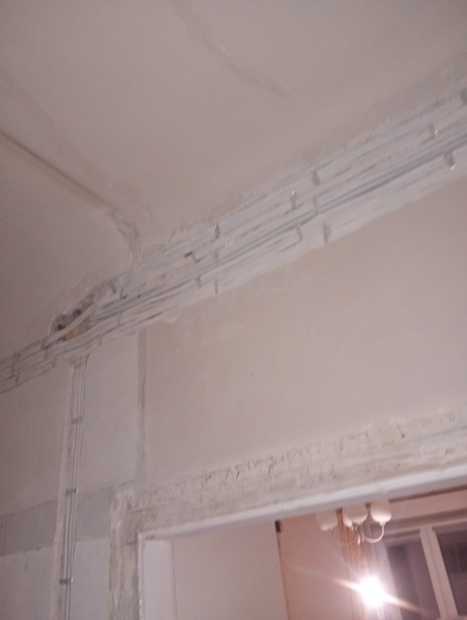 Białe rurki instalacji elektrycznej, przymocowane do sufitu i ściany w surowym stanie, widoczne przed tynkowaniem. Fragment widoku przez otwór drzwiowy.