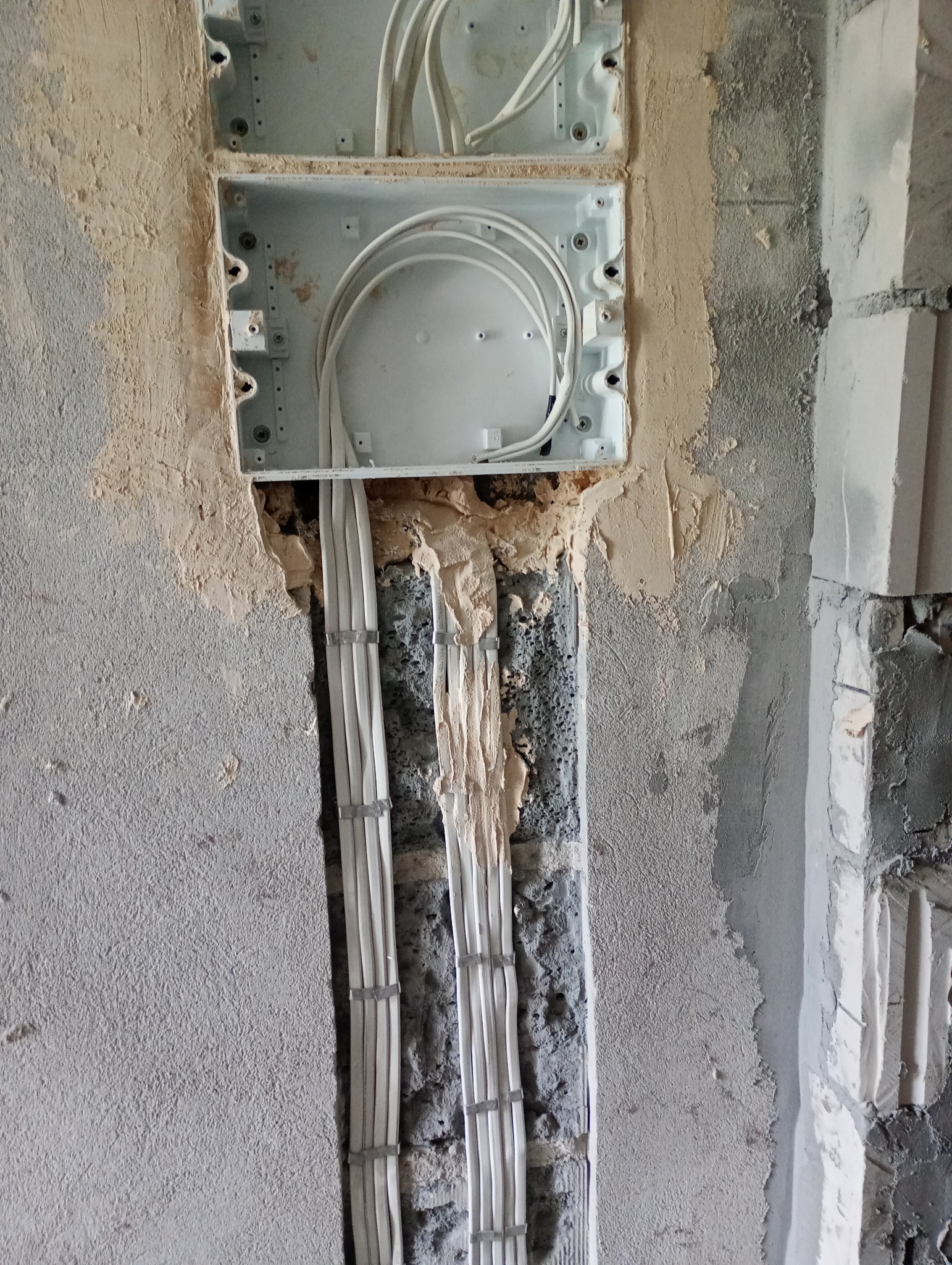 Białe przewody elektryczne prowadzone w ścianie, zamocowane w metalowej puszce instalacyjnej. Widoczne surowe wykończenie ściany i przewody zabezpieczone gipsem.