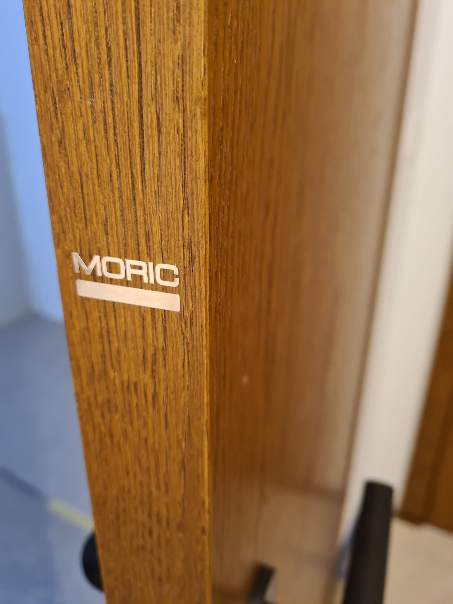 Fragment drewnianych drzwi z widocznym srebrnym logo producenta 'MORIC' i teksturą drewna.