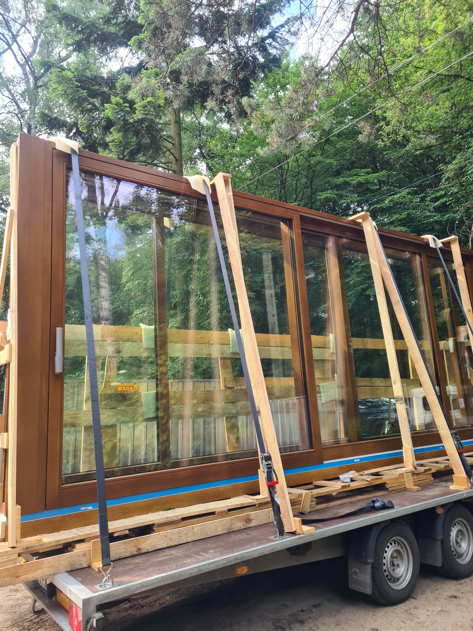 Duże, brązowe okno przesuwne z szybami, zabezpieczone drewnianymi elementami i czarnymi pasami transportowymi, umieszczone na przyczepie samochodowej na tle zieleni.