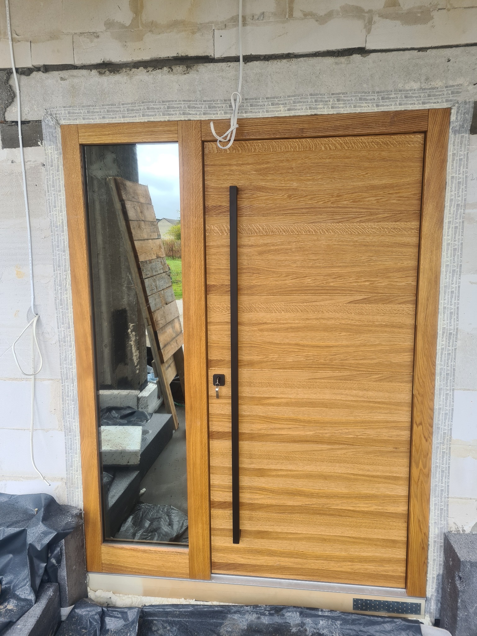 Nowoczesne drzwi wejściowe z drewnianą okładziną i długim, pionowym uchwytem, z przeszklonym bocznym panelem, zamontowane w surowym stanie budynku z widocznymi bloczkami i izolacją.