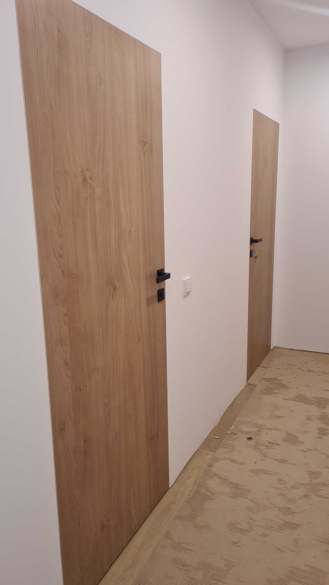 Dwa skrzydła drzwi wewnętrznych o drewnopodobnym wykończeniu i czarnych klamkach, widoczne w nowym budynku z niedokończoną podłogą.