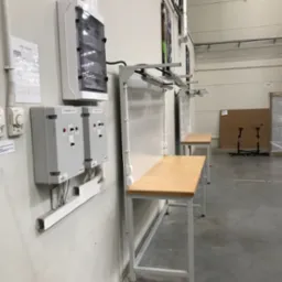 Szare skrzynki sterownicze z przewodami na ścianie w hali produkcyjnej obok stołów roboczych z metalowymi nogami i drewnianymi blatami.