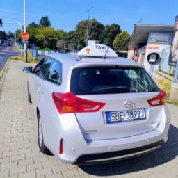 Srebrny samochód typu kombi z oznaczeniem taxi na dachu, zaparkowany na chodniku. Widoczna tablica rejestracyjna, znak STOP i budka telefoniczna w tle.