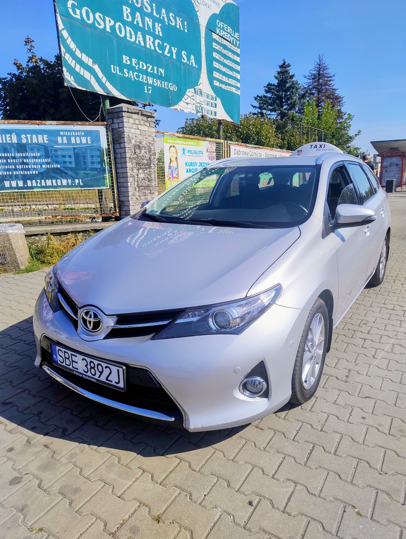 Srebrny samochód typu kombi z oznaczeniem TAXI na dachu, zaparkowany na chodniku z kostki brukowej, z billboardami w tle.
