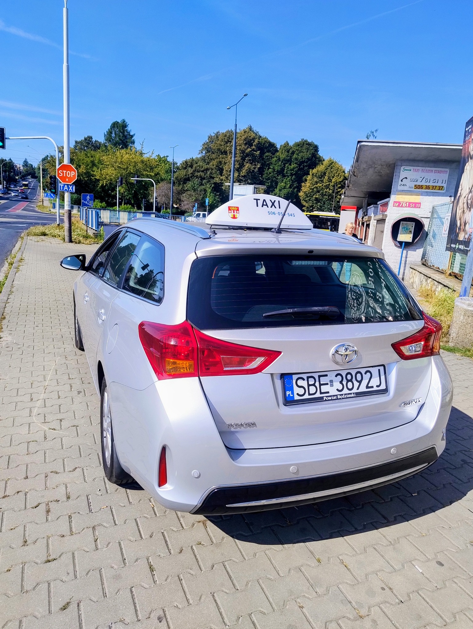 Srebrny samochód typu kombi z oznaczeniem taxi na dachu, zaparkowany na chodniku. Widoczna tablica rejestracyjna, znak STOP i budka telefoniczna w tle.