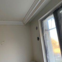 Przemek - Wnętrze po remoncie: świeżo pomalowane ściany w odcieniach bieli, dekoracyjny gzyms sufitowy, okno zabezpieczone folią malarską i widoczne otwory pod instalacje.