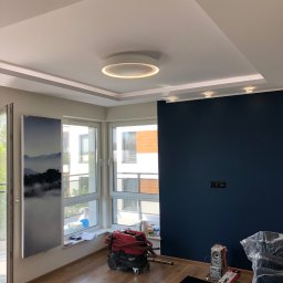 Przemek - Nowoczesne wnętrze po remoncie w Golub-Dobrzyń: ciemna ściana, podwieszany sufit z oświetleniem LED, okna z widokiem na zewnątrz. Na podłodze widoczny sprzęt budowlany.