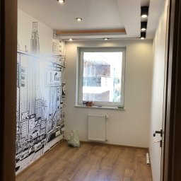 Przemek - Pokój po remoncie: drewniana podłoga, białe ściany z tapetą w stylu miejskim, okno z grzejnikiem, nowoczesne oświetlenie sufitowe.