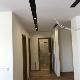 Przemek - Jasne wnętrze z drewnianą podłogą i drzwiami. Sufit z czarnymi listwami oświetleniowymi. Widoczne świeżo pomalowane ściany i wystające przewody.