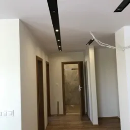 Jasne wnętrze z drewnianą podłogą i drzwiami. Sufit z czarnymi listwami oświetleniowymi. Widoczne świeżo pomalowane ściany i wystające przewody.