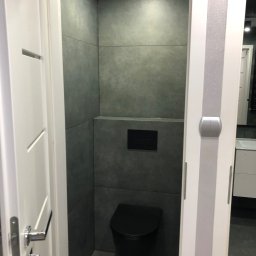 Przemek - Nowoczesna toaleta z szarymi płytkami, czarną miską ustępową i białymi drzwiami z srebrnymi akcentami. Widok zza otwartych drzwi.