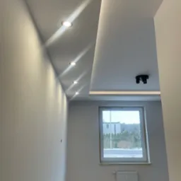 Nowoczesne wnętrze: białe ściany, sufit podwieszany z oświetleniem punktowym, okno z widokiem na zewnątrz, grzejnik. Minimalizm i jasne barwy.