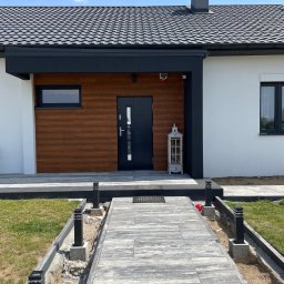 Przemek - Nowoczesny dom jednorodzinny z drewnianą elewacją przy wejściu i szarym chodnikiem z kostki. Widoczny fragment dachu z kominem i oświetlenie zewnętrzne.