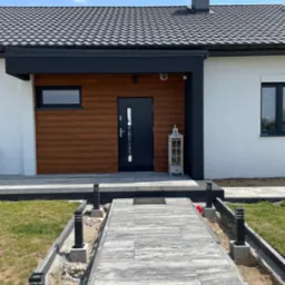 Nowoczesny dom jednorodzinny z drewnianą elewacją przy wejściu i szarym chodnikiem z kostki. Widoczny fragment dachu z kominem i oświetlenie zewnętrzne.