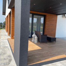 Przemek - Nowoczesny taras z drewnianą podłogą i elewacją, szary filar z fakturą, meble ogrodowe i lampiony. Widok na zadbany ogród w tle.