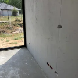 Szara ściana z wywierconymi otworami na gniazdka elektryczne, surowy beton na podłodze, widok na zewnątrz przez duże okno. Wnętrze w trakcie wykańczania.