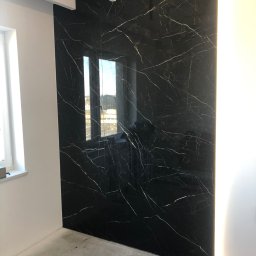 Przemek - Ściana wykończona czarnymi płytami imitującymi marmur z białymi żyłami, podświetlona LED w Golub-Dobrzyń. Widoczne okno i sufit z oświetleniem.