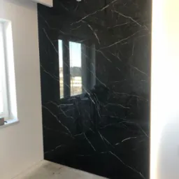 Ściana wykończona czarnymi płytami imitującymi marmur z białymi żyłami, podświetlona LED w Golub-Dobrzyń. Widoczne okno i sufit z oświetleniem.