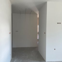 Przemek - Jasny korytarz z gładkimi, białymi ścianami i surową betonową posadzką, z widocznymi otworami pod gniazdka oraz zwisającymi przewodami oświetleniowymi.