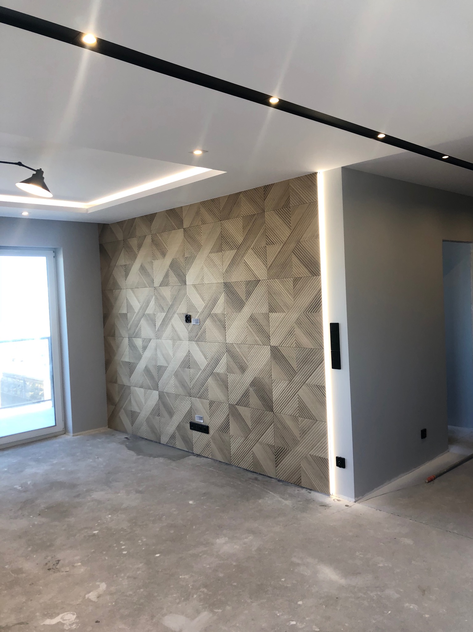 Nowoczesne wnętrze: ściana z geometrycznym wzorem, podświetlenie LED, szara ściana, surowa podłoga, listwy oświetleniowe. Włączniki na ścianie.