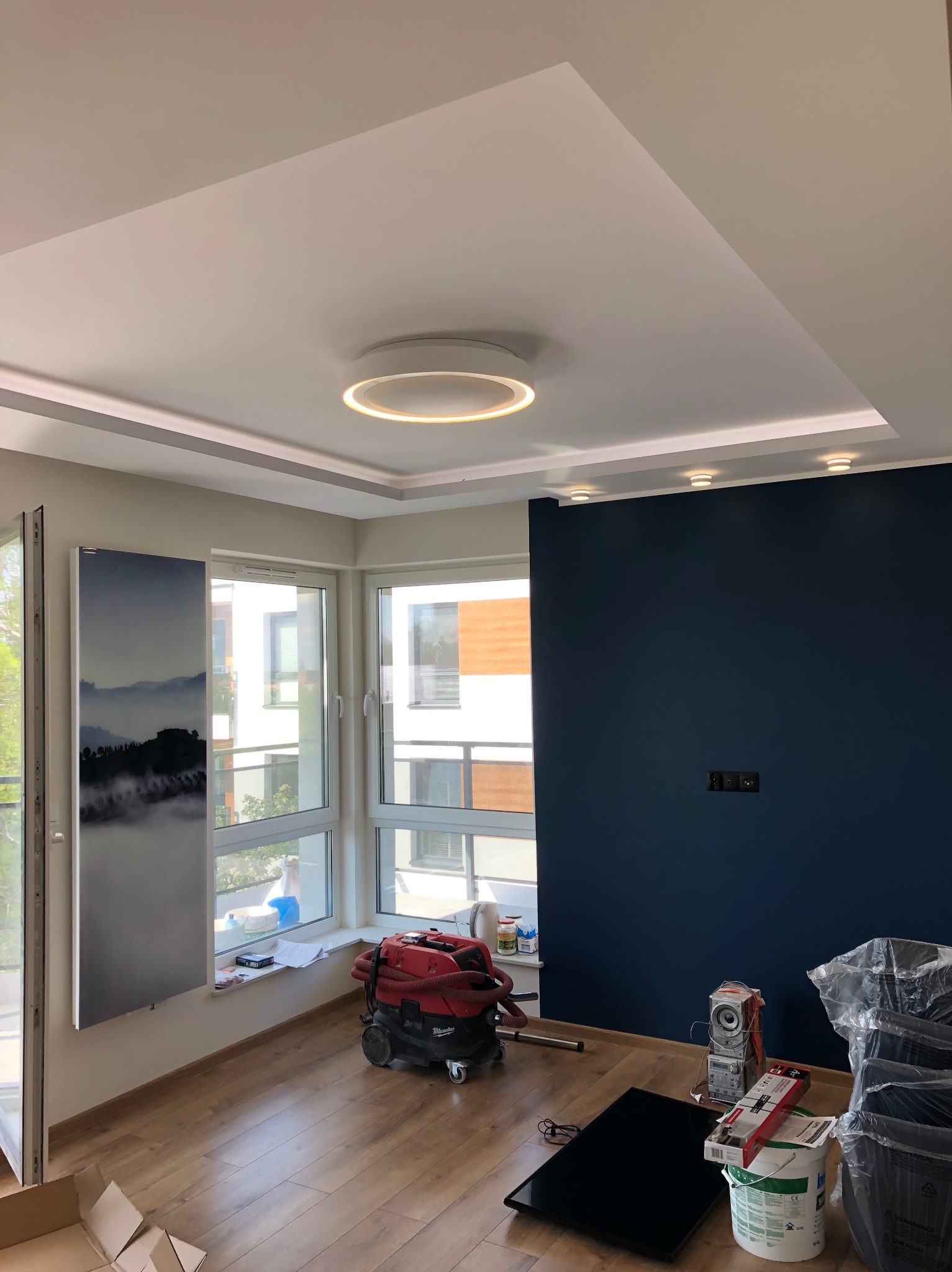 Nowoczesne wnętrze po remoncie w Golub-Dobrzyń: ciemna ściana, podwieszany sufit z oświetleniem LED, okna z widokiem na zewnątrz. Na podłodze widoczny sprzęt budowlany.